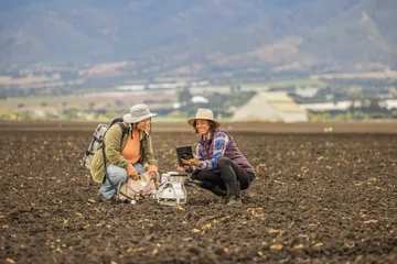 A professor and her research assistant work in a farm field, examining data on a tablet next to scientific equipment and soil sensors.