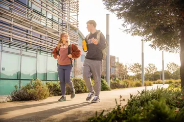 Two CSUMB college students walk and chat on a sunny campus path, carrying backpacks and a water bottle near a modern building.