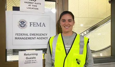 Alora Skustad stands next to three signs: Sign 1: No hazmat suits at this location; Sign 2: FEMA, Federal Emergency Management System; Sign 3: Returning guests please go to garage Level 1.