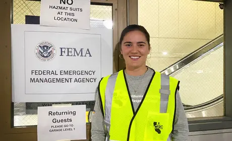 Alora Skustad stands next to three signs: Sign 1: No hazmat suits at this location; Sign 2: FEMA, Federal Emergency Management System; Sign 3: Returning guests please go to garage Level 1.