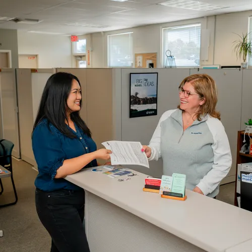 A CSUMB employee with long black hair hands a document to an employee with short blond hair standing behind a reception counter in a bright, simple office.