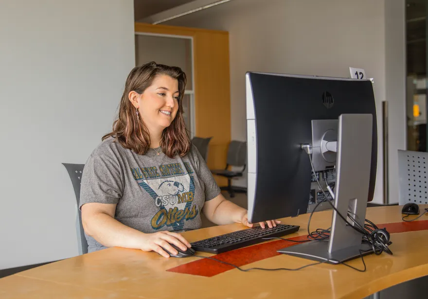 A student with blond hair wearing a CSUMB Otters T-shirt sits in front of a computer monitor smiling.
