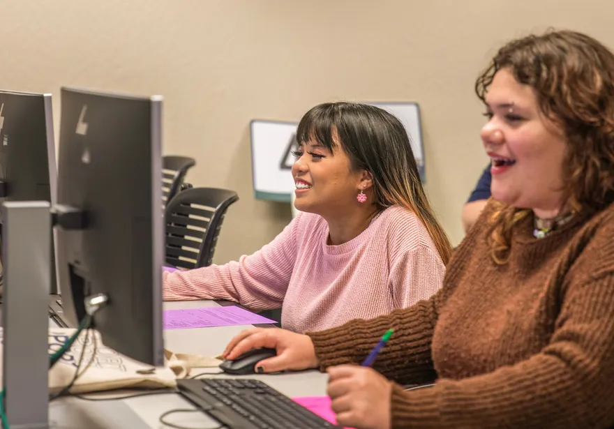 Two students smiling and looking at desktop computers